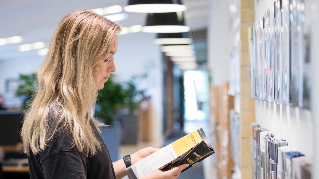 An image slider with an image of a woman looking at a brochure while standing in front of a brochure wall.